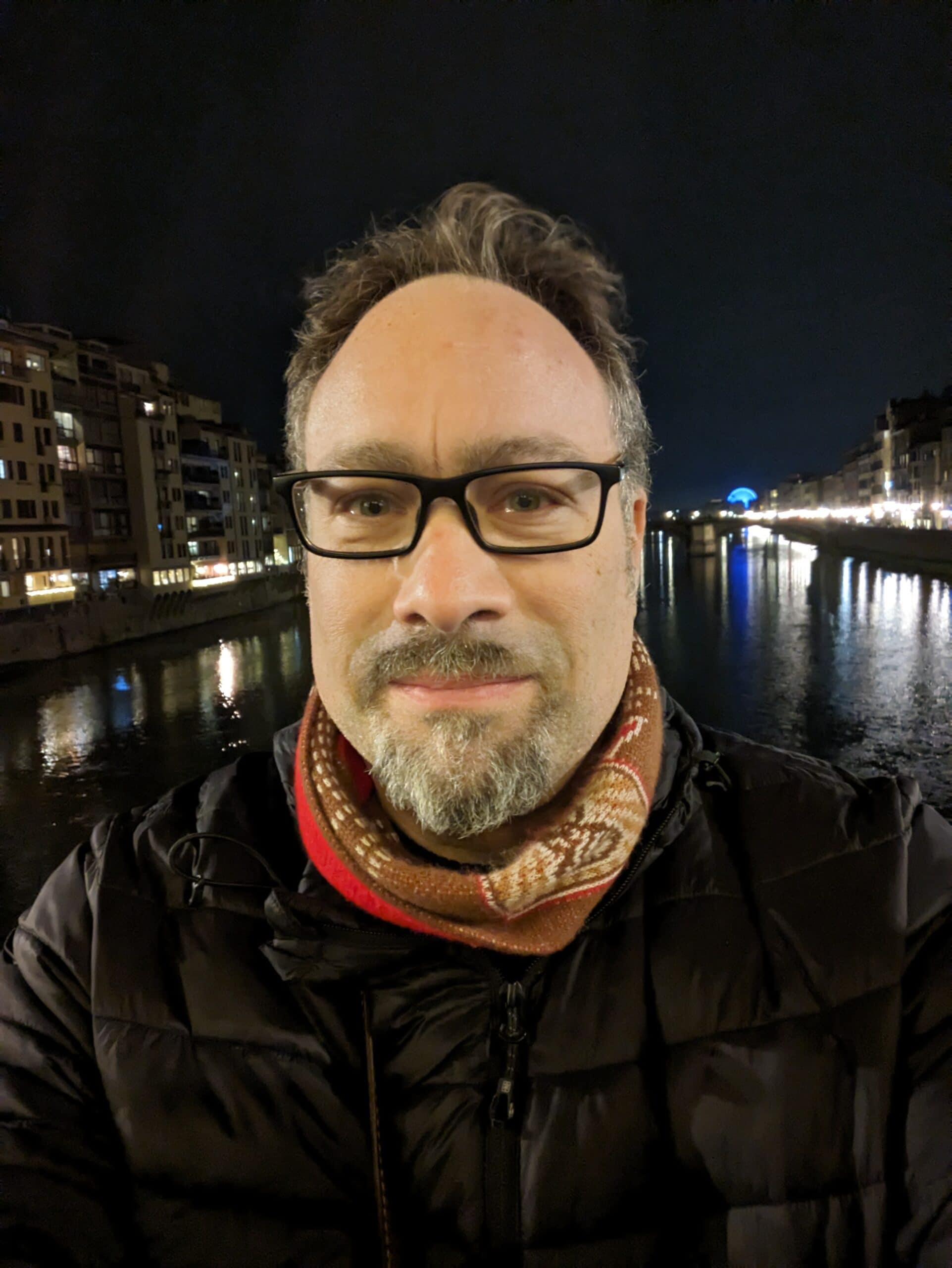 A man taking a nighttime selfie along a city canal with illuminated buildings, reflecting water, and a distinctive Ferris wheel in the distance, showcasing urban nightlife and scenic views in Italy.
