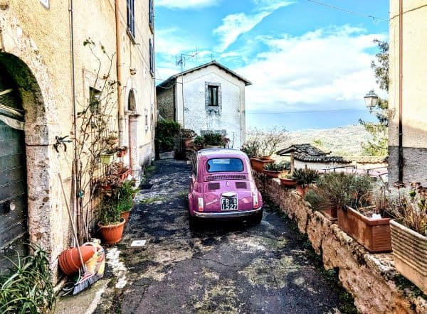 Vintage pink Fiat 500 parked on a narrow rustic street in an Italian village, with flowering potted plants and charming stone buildings, showcasing authentic Italian countryside scenery.