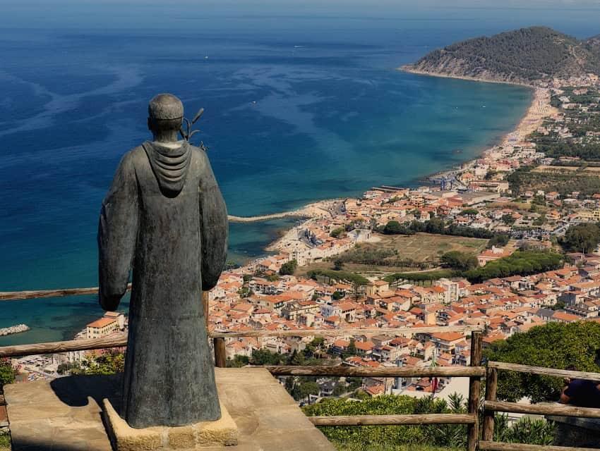 A bronze statue of a monk gazes over a picturesque Italian seaside town with colorful buildings, sandy beaches, and turquoise waters, capturing the beauty of Italy’s coastal landscapes.