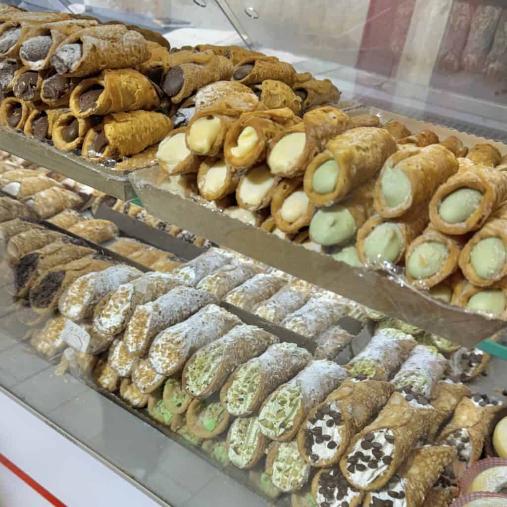 A variety of traditional Italian pastries including cannoli filled with creamy ricotta, decorated with chocolate chips and powdered sugar, showcased in a bakery display case.
