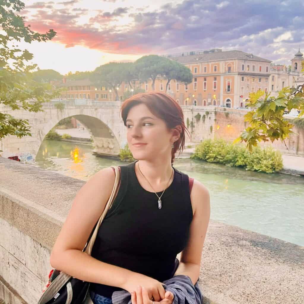 Young woman enjoying a sunset by the Tiber River in Rome, with historic bridges and cityscape in the background, capturing the essence of Italy's timeless beauty.