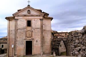Ancient stone church with a cross on top, set against a cloudy sky, surrounded by ruins and old buildings in Italy.