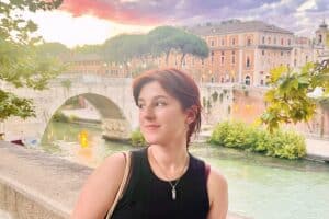 Young woman with short red hair standing by the Tiber River in Rome, Italy, during sunset, with historic buildings and a stone bridge in the background.