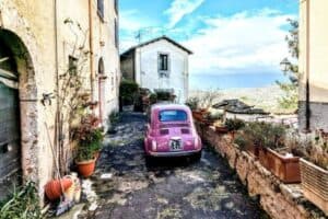 A picturesque narrow alley in Italy featuring a pink vintage car parked on a rustic stone path, surrounded by potted plants and old buildings with scenic views in the background.