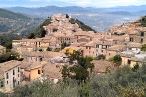 Panoramic view of a traditional Italian hillside village with historic stone buildings and terracotta roofs, surrounded by lush greenery and rolling hills in the background.