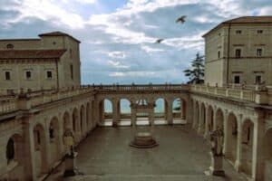 Ancient Italian palace with classical architecture overlooking the sea, featuring arched windows and statues, under a partly cloudy sky with flying birds.