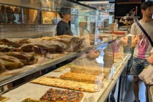 Freshly baked pizzas and traditional Italian dishes showcased in a busy pizzeria with customers ordering and staff preparing food behind the glass counter.