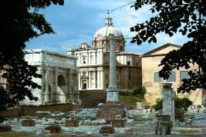 Historical Roman Forum ruins with ancient columns and structures in Rome, Italy, under a blue sky with trees framing the scene.