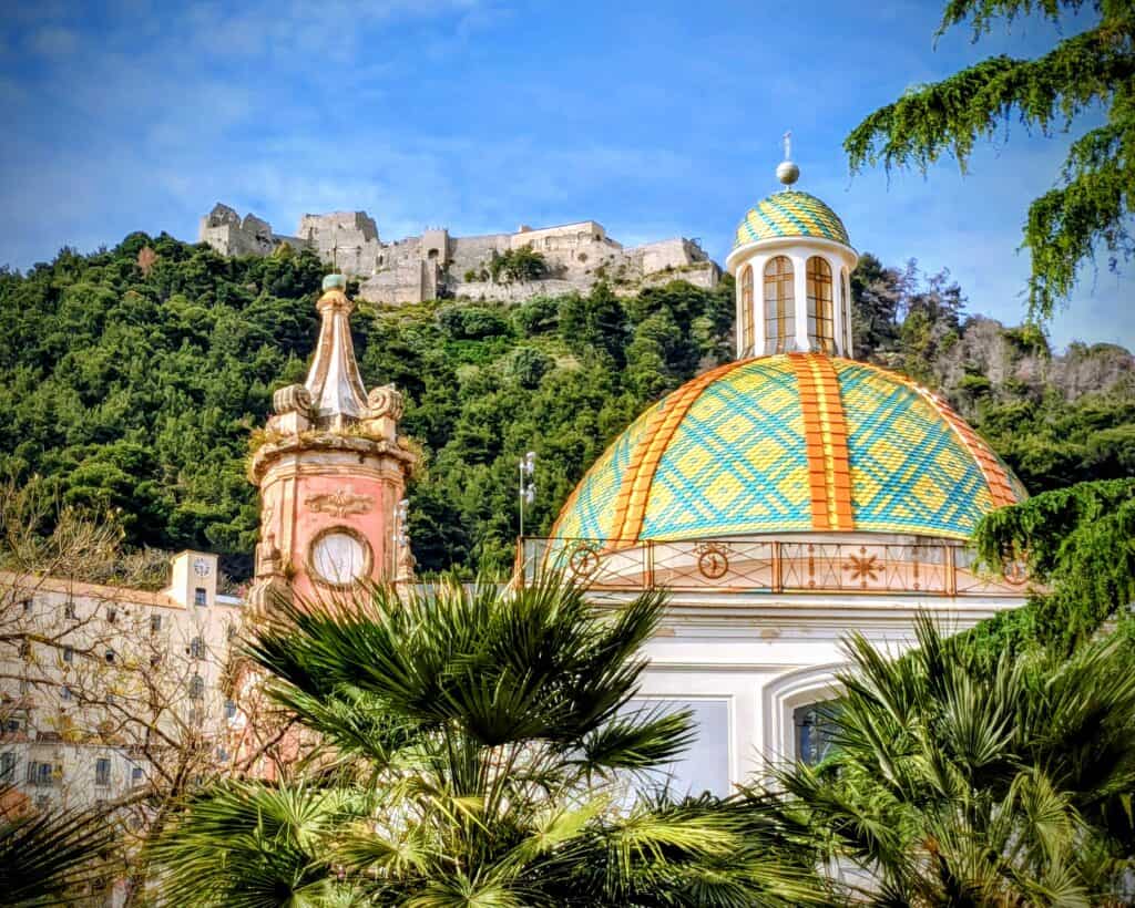 Vibrant colorful dome with historic castle on a lush hillside in Italy, under a bright blue sky, surrounded by green trees and tropical plants.