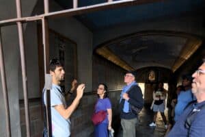 Group of tourists listening to a guide explaining history inside a historic site in Italy, with a barred window in the foreground.